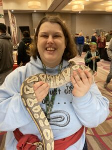 Amber smiles broadly and holds a large python. The snake is patterned in varying shades of brown and is curled around Amber’s hands.