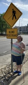 Janeen wears a shirt with horizontal rainbow stripes and holds a white cane. She stands on a sidewalk under a sign warning of blind pedestrians.