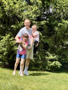 Matt Selm holding his youngest daughter. His older daughter is about chest height in front of him. 