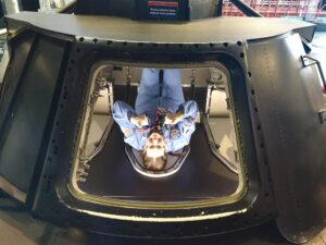 The image shows Melanie lying inside a replica Apollo spacecraft module at the US Space and Rocket Center in Huntsville, Alabama. She is wearing a blue flight suit with various patches and is giving a thumbs-up. The module is black with a large, square opening through which she is visible inside the metallic interior.