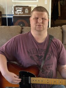 Justin sitting on a couch, holding an electric guitar. He is wearing a maroon T-shirt with the word "MEMPHIS" and a graphic of a guitar printed on it.