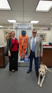 Melanie Alt Sinohui, Ron Brooks, and Ron’s guide dog, York, standing in front of Mark Kelly's flight suit in his office on Capitol Hill