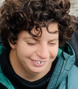 Sara Mornis wearing a dark top with a green outdoor-style jacket over it, which has a hood and a textured, slightly shiny fabric. Sara has dark brown curly hair.