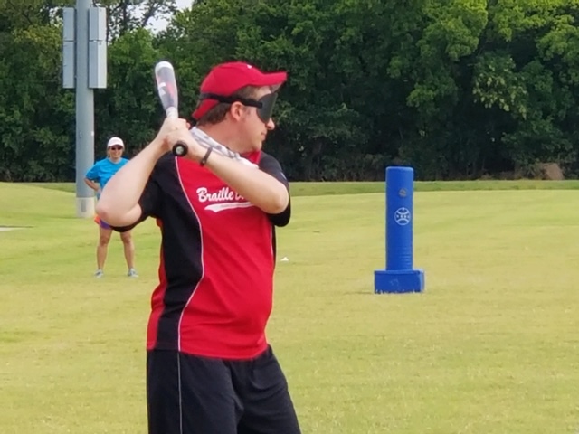 A photo of Greg Lindberg holding a bat over his shoulder; he is wearing his Braille Bandits jersey which is red and black and a Braille Bandits hat which is red; the photo is from the 2019 National Beep Baseball Association World Series in Tulsa, OK
