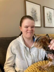 Vika sits on a couch and holds a small dog with long brown and black fur. She wears a zip-up hoodie with her hair tied back. There are two pieces of abstract art on the wall behind her.