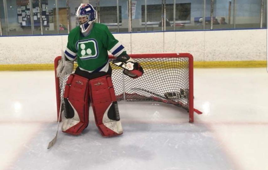 Liz standing in front of a hockey net. She is wearing a green jersey with the number 30, along with red pads, gloves, and a helmet